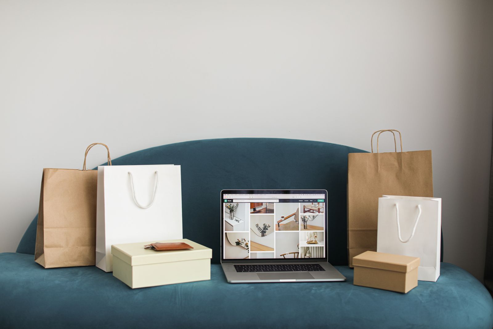 A laptop showing a grid of product photos, surrounded by paper shopping bags and gift boxes on a sofa.